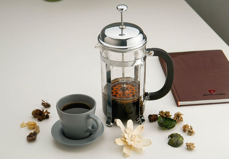 A glass French press with a cup of coffee on a light table